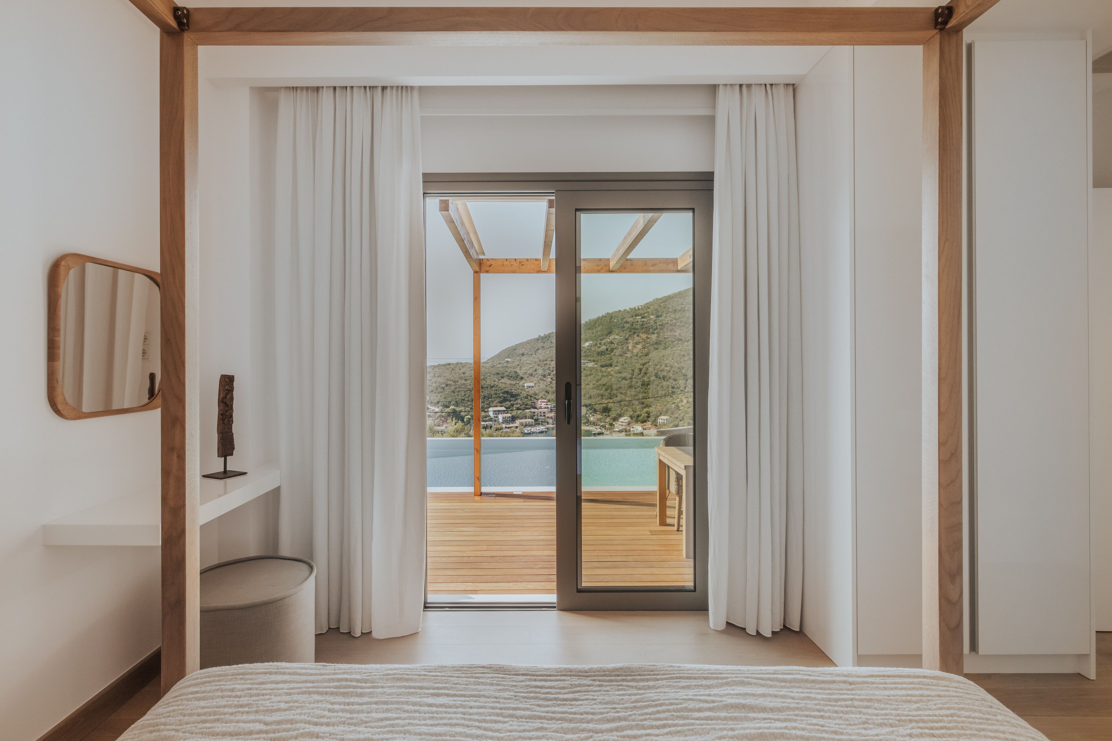 A bedroom with a large bed, a wooden ceiling, a sliding glass door to a balcony, and a view of a hillside.