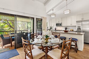 A modern kitchen with white cabinets, stainless steel appliances, and a glass dining table surrounded by wicker chairs. - Relax in Style, Tee Off in Paradise (Kihei)