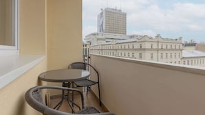 Un balcón con una mesa redonda y dos sillas de mimbre, con vistas a un paisaje urbano que incluye edificios históricos y una estructura moderna alta.