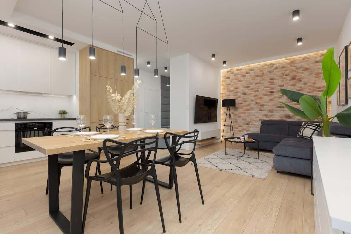 Une cuisine moderne avec des meubles en bois, un coin repas avec une table en bois et des chaises noires, un canapé gris et un mur d’accent en briques.