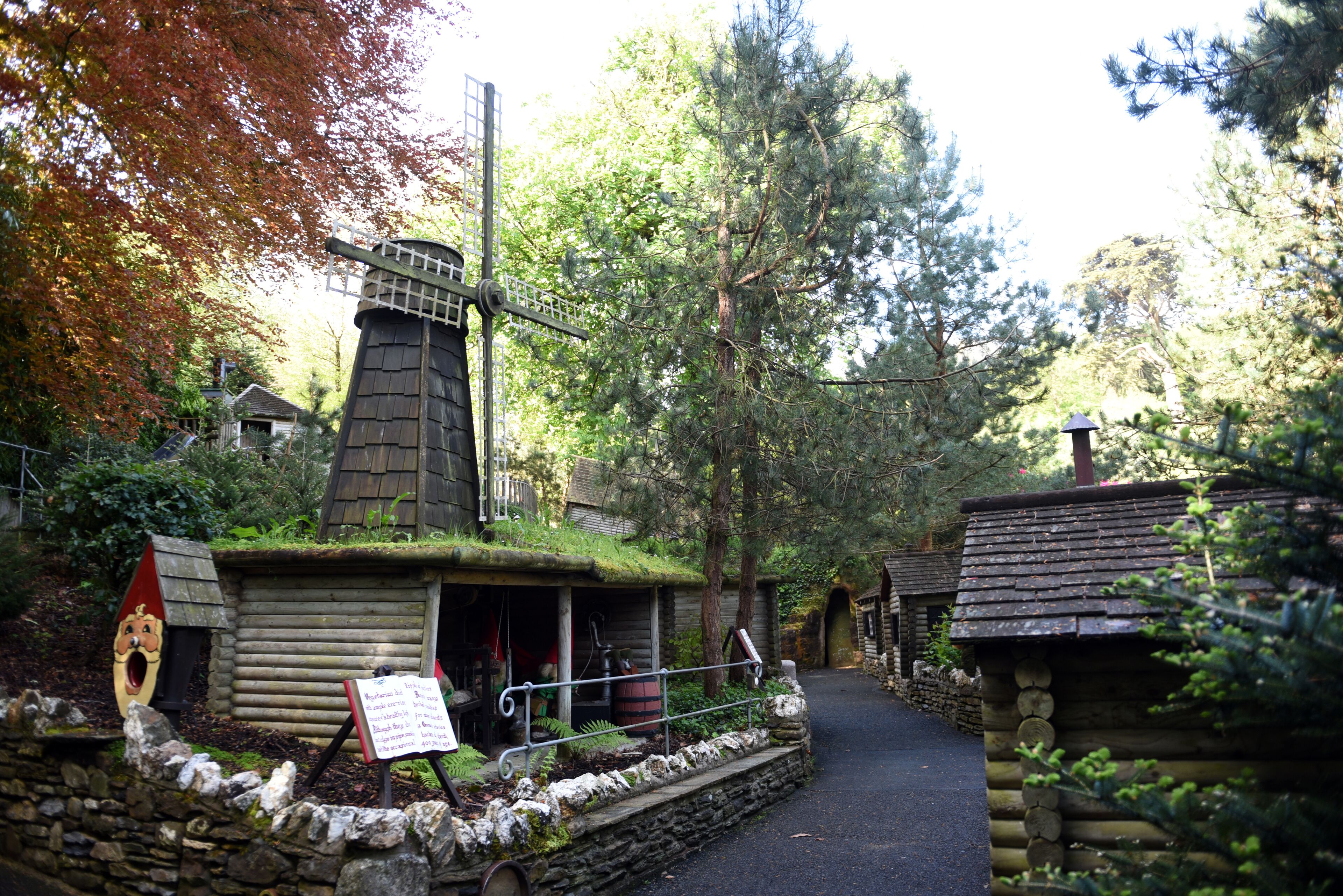 A rustic log cabin with a windmill, surrounded by trees and a stone wall.