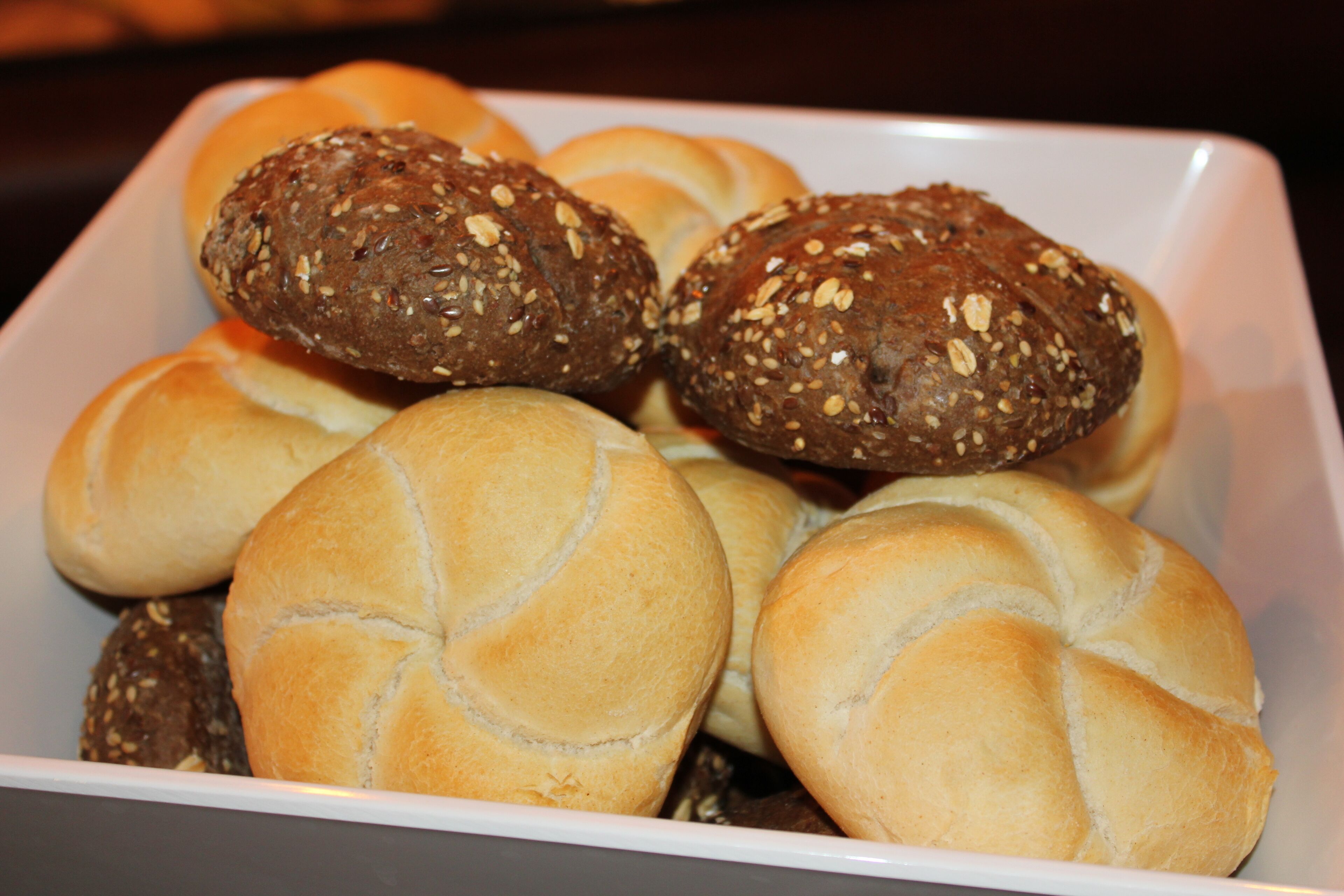 A tray of assorted bread rolls, including sesame and seed varieties, arranged on a white plate.