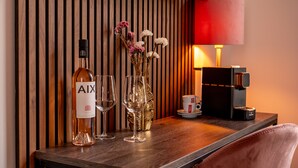 Una mesa de madera con una botella de vino AIX, dos copas de vino, un florero con flores, una cafetera y una lámpara roja.