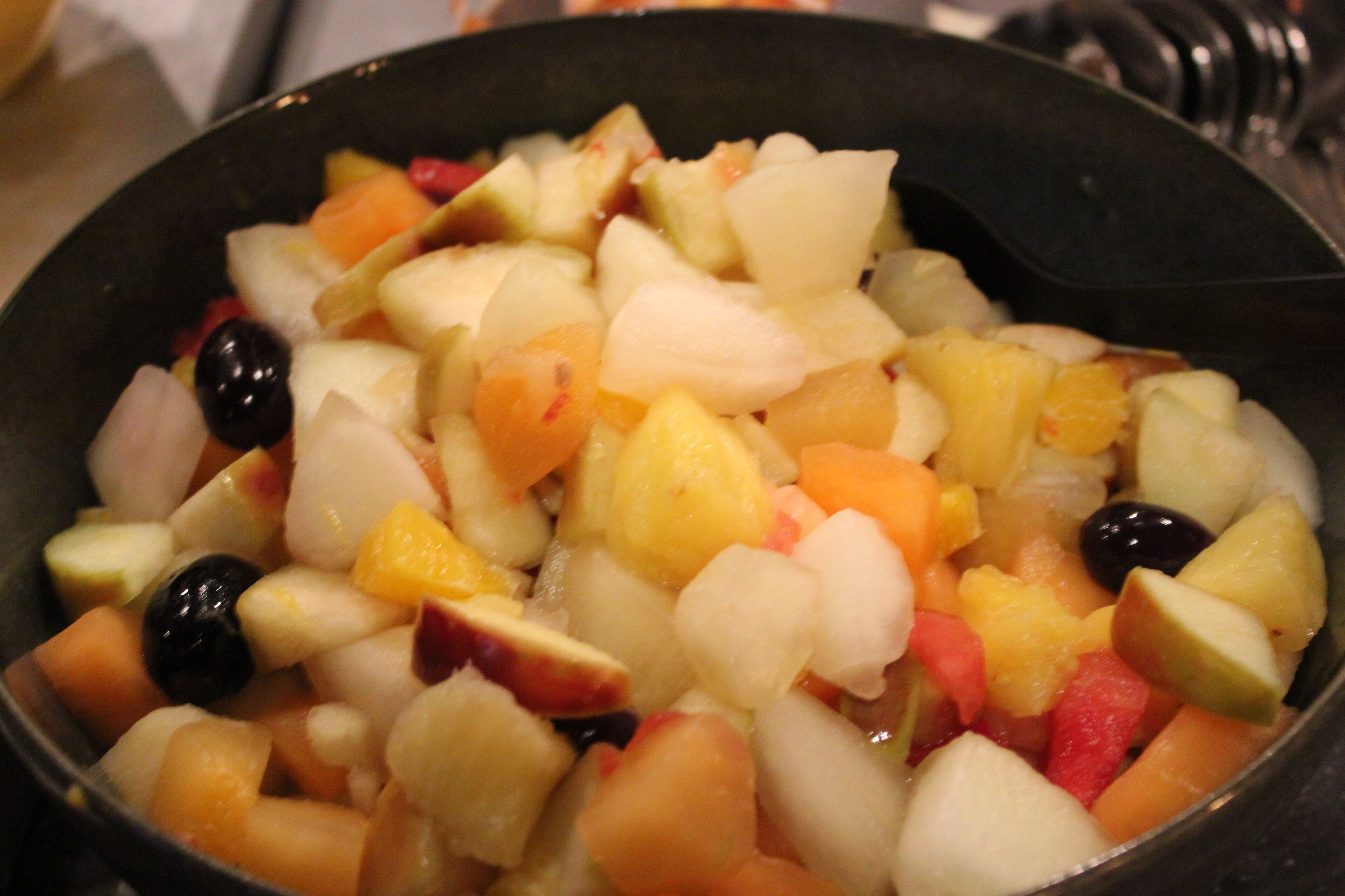A bowl of mixed fruit salad with various fruits and a black spoon.