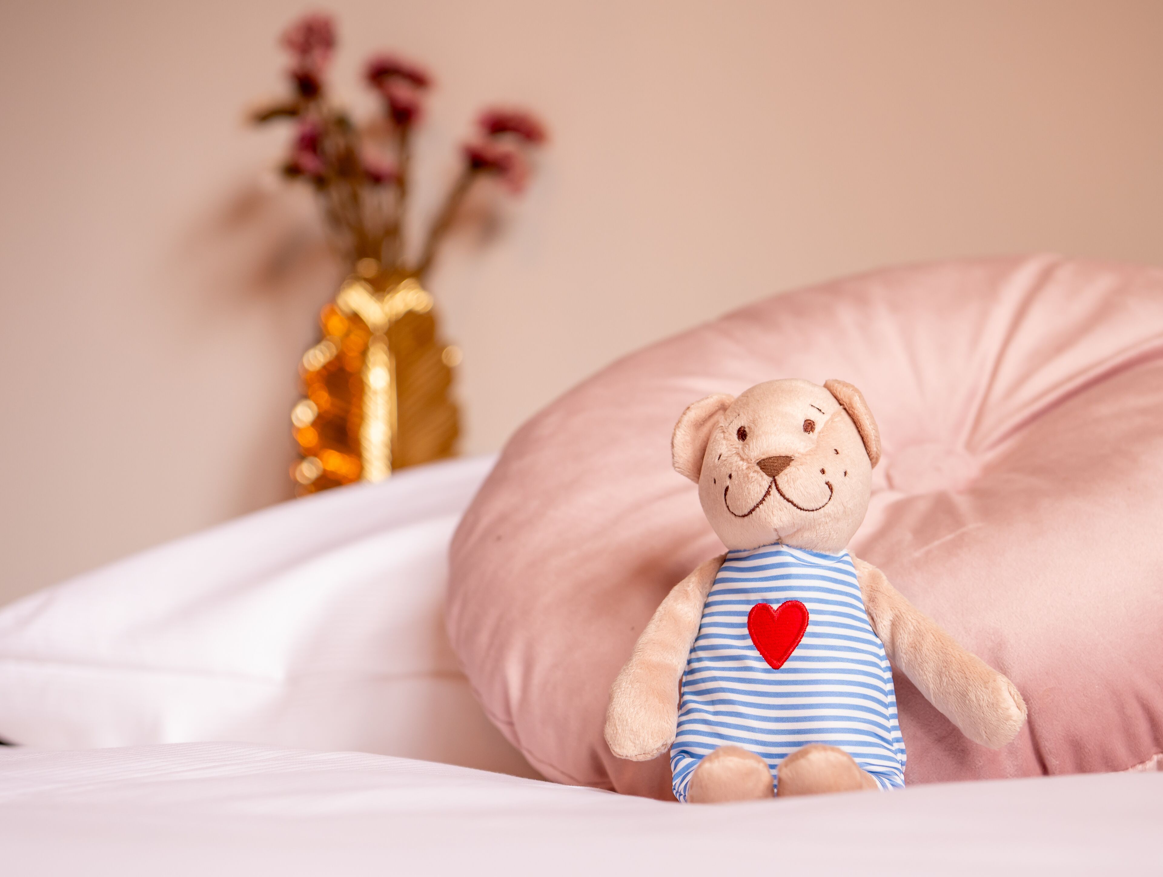 Un peluche con un corazón en el pecho, sobre una cama con una almohada rosa y un jarrón con flores al fondo.