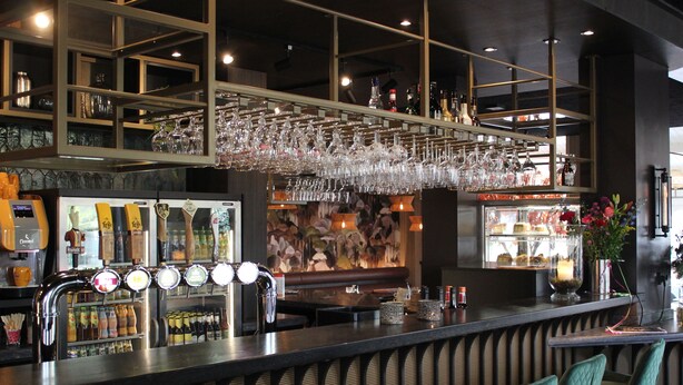 Zona de bar con mostrador, múltiples grifos y vasos colgando. Hay sillas y un jarrón decorativo con flores.