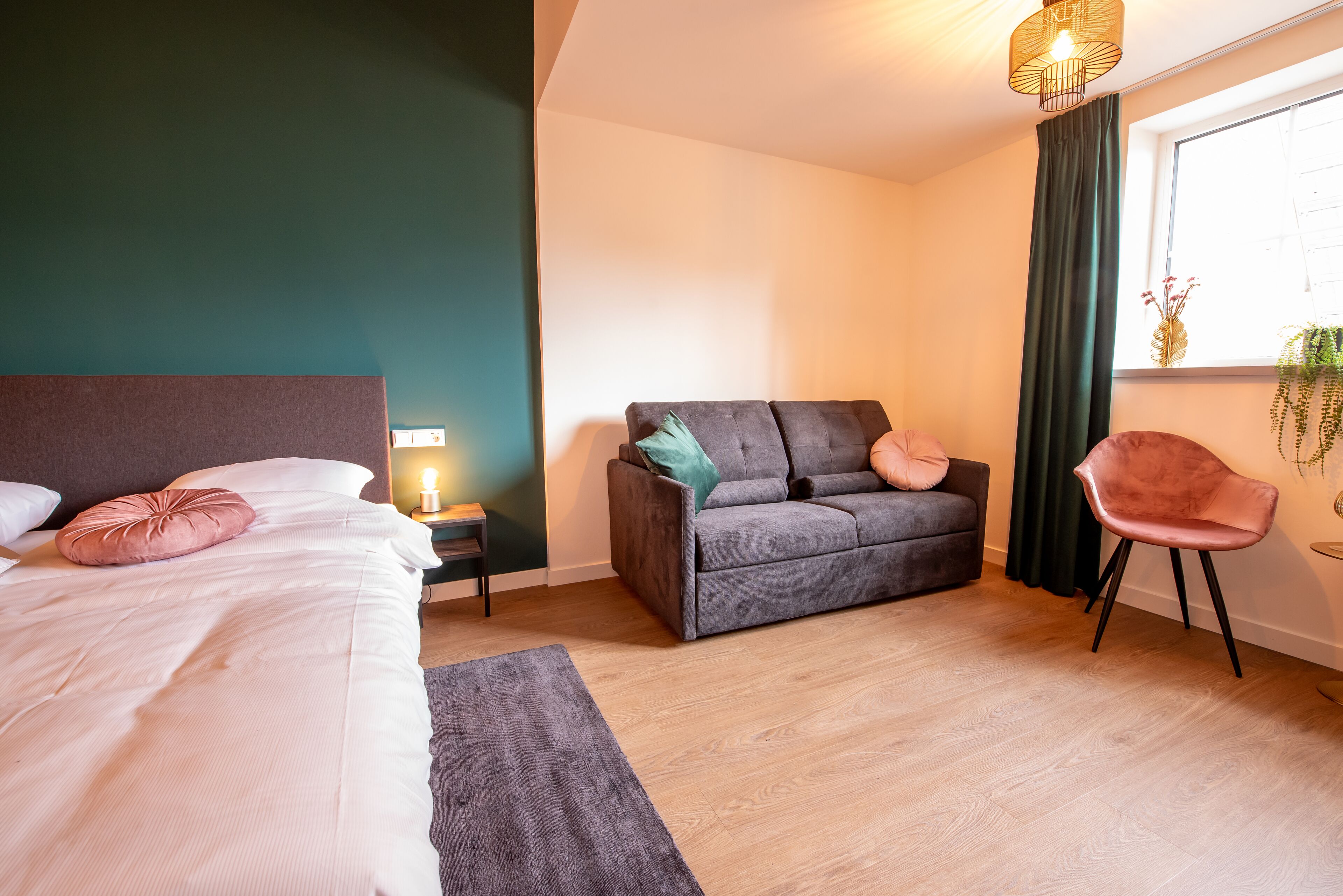 A modern bedroom with a bed, a grey sofa, a pink chair, and a small table with a lamp.