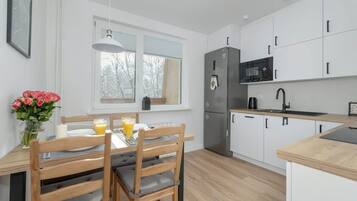 Una cocina moderna con armarios blancos, un refrigerador de acero inoxidable, un microondas y un lavamanos. Hay una mesa de comedor con sillas y un florero con flores.