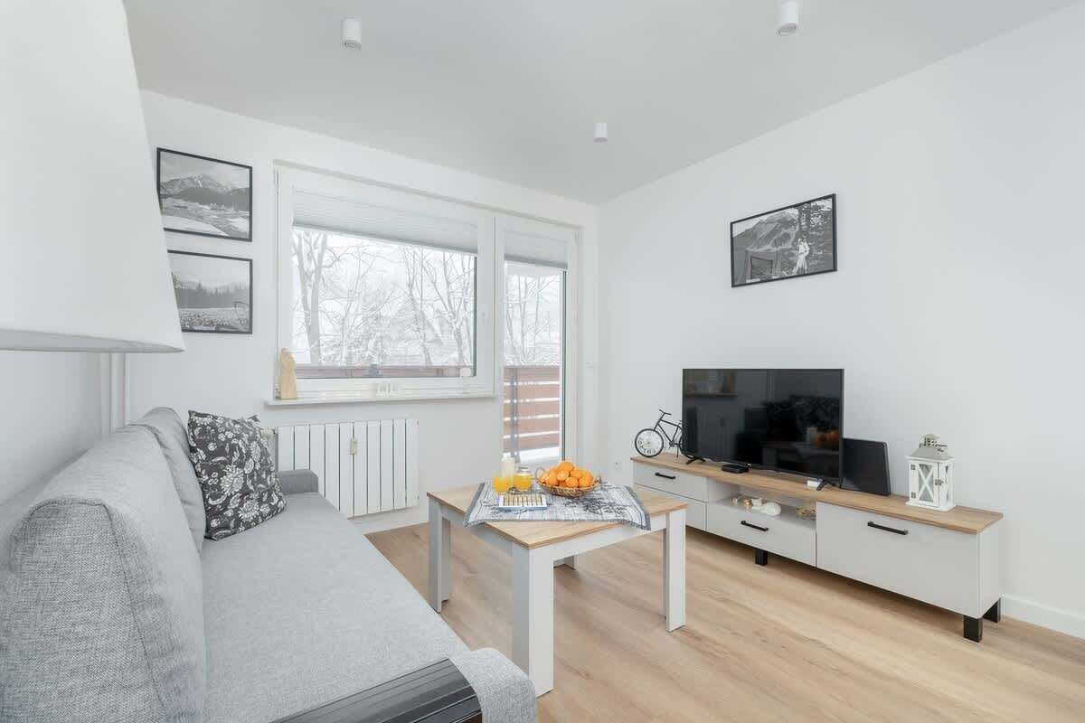 Una sala moderna con un sofá gris, una mesa de centro de madera con frutas y un periódico, un televisor sobre un mueble blanco, y una ventana con vista a árboles.