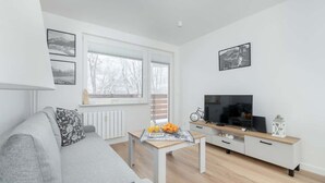 A modern living room with a grey sofa, a wooden coffee table with fruits and a newspaper, a TV on a white cabinet, and a window with a view of trees. - Apartment with a View of the Tatra Mountains | Balcony | 600 m to the Center (Zakopane)