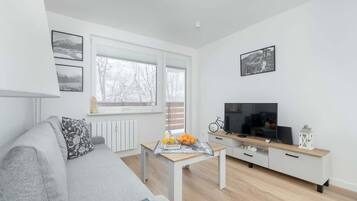 Una sala moderna con un sofá gris, una mesa de centro de madera con frutas y un periódico, un televisor sobre un mueble blanco, y una ventana con vista a árboles.