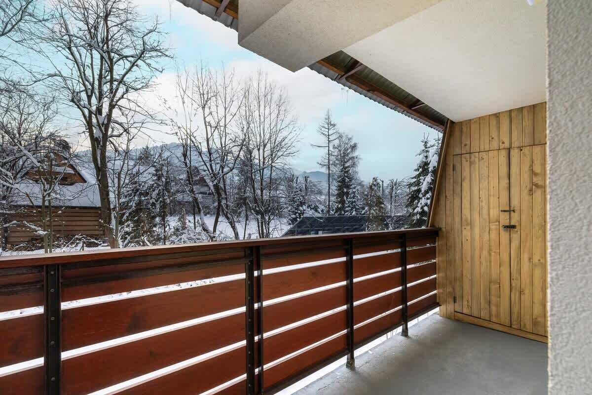 Un balcón con barandilla de madera que da a un paisaje nevado con árboles y un edificio al fondo.