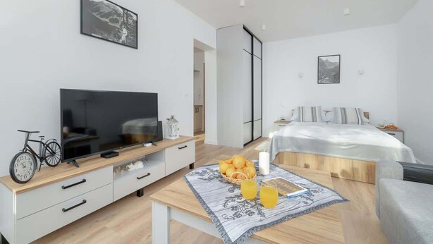 A modern hotel room with a flat-screen TV, a dining table with fruit and drinks, a bed with a white duvet, and a large window. - Apartment with a View of the Tatra Mountains | Balcony | 600 m to the Center (Zakopane)