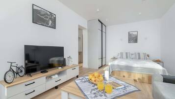 Una habitación de hotel moderna con una televisión de pantalla plana, una mesa de comedor con fruta y bebidas, una cama con edredón blanco y un ventanal grande.