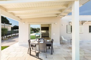 An outdoor patio with a table and chairs, a covered area, and a view of a swimming pool and greenery. - 3642 Villa Angi by Perle di Puglia (San Vito dei Normanni)