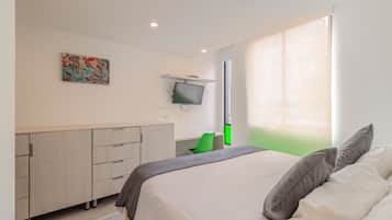 Un dormitorio con cama, televisor montado en la pared, escritorio y ventana con cortinas.