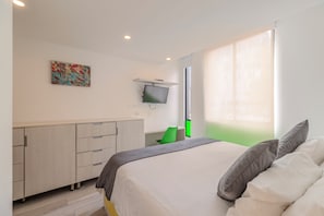 Une chambre à coucher avec un lit, un téléviseur fixé au mur, un bureau et une fenêtre avec des rideaux.