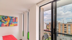 A room with a bed, a colorful abstract painting, a window with a view of buildings, and a chair. - DAS HAUS  (Bogotá)