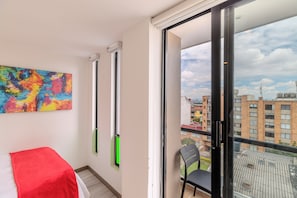 Ein Zimmer mit einem Bett, einem farbenfrohen abstrakten Gemälde, einem Fenster mit Blick auf Gebäude und einem Sessel.
