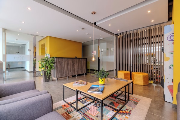 Un área de recepción moderna con un mostrador de madera, un tapete colorido y un espacio de descanso con dos otomanas.