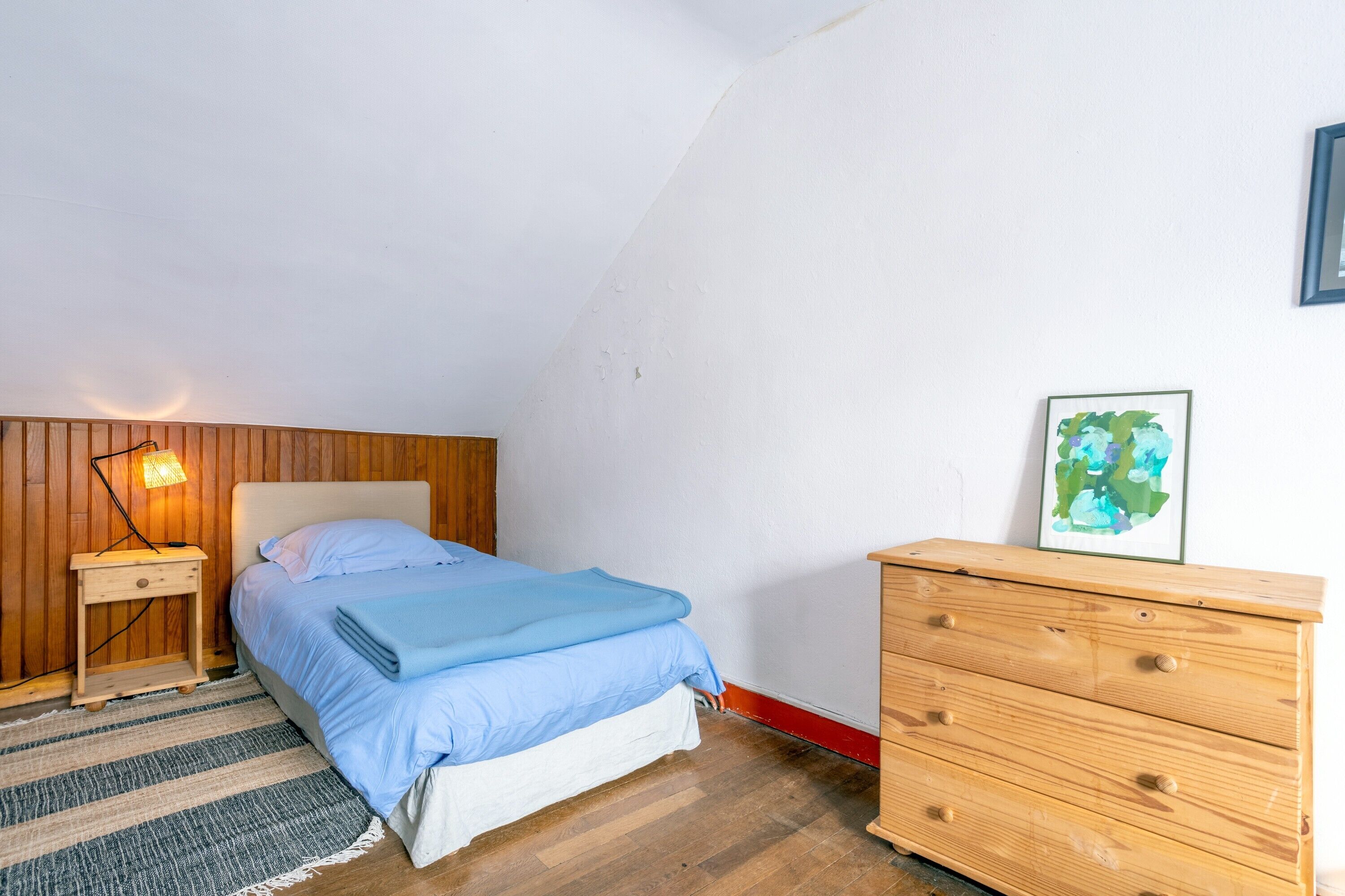 A bedroom with a bed, a nightstand, a wooden dresser, a wall-mounted lamp, and a framed picture.