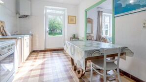 A kitchen with a tiled floor, a dining table with chairs, a window with a view of greenery, and a wall-mounted mirror. - Small House 5min Drive from Beach with Private Terrace and Garden (Plobannalec-Lesconil)