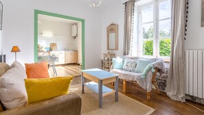 A living room with a sofa, a table, a chair, a lamp, a mirror, and a view of the kitchen. - Small House 5min Drive from Beach with Private Terrace and Garden (Plobannalec-Lesconil)