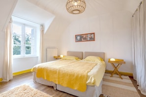 Une chambre avec un grand lit, deux tables de chevet, une fenêtre avec des rideaux, un luminaire au plafond et un tableau encadré au mur.