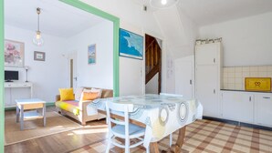 A room with a dining table, a sofa with cushions, a television, and a kitchen area with cabinets. - Small House 5min Drive from Beach with Private Terrace and Garden (Plobannalec-Lesconil)