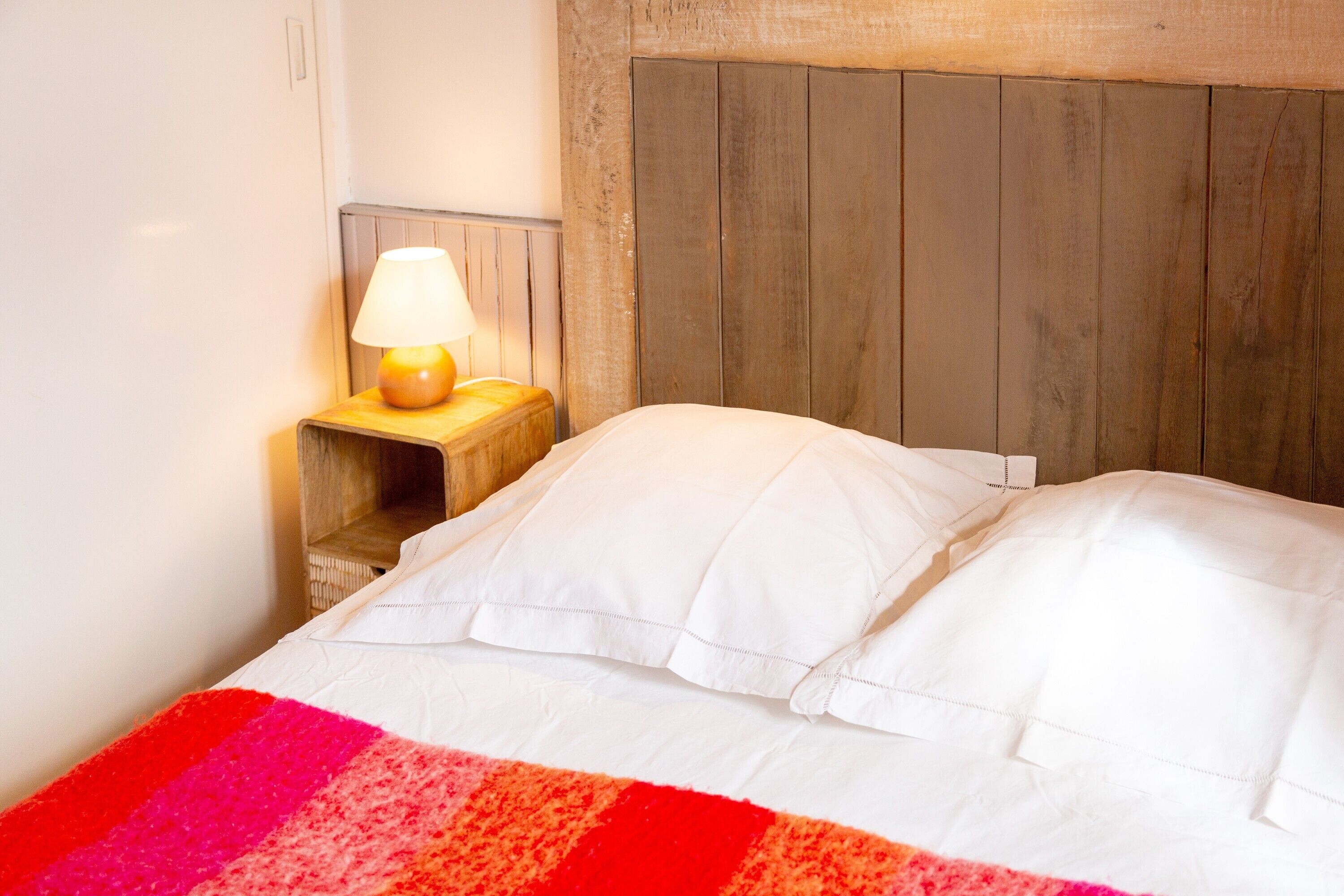 A bed with white pillows and a multicolored blanket, a wooden headboard, and a nightstand with a lamp.