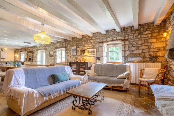 A cozy living room with stone walls, wooden beams, a sofa, armchairs, a coffee table, and a hanging light fixture.