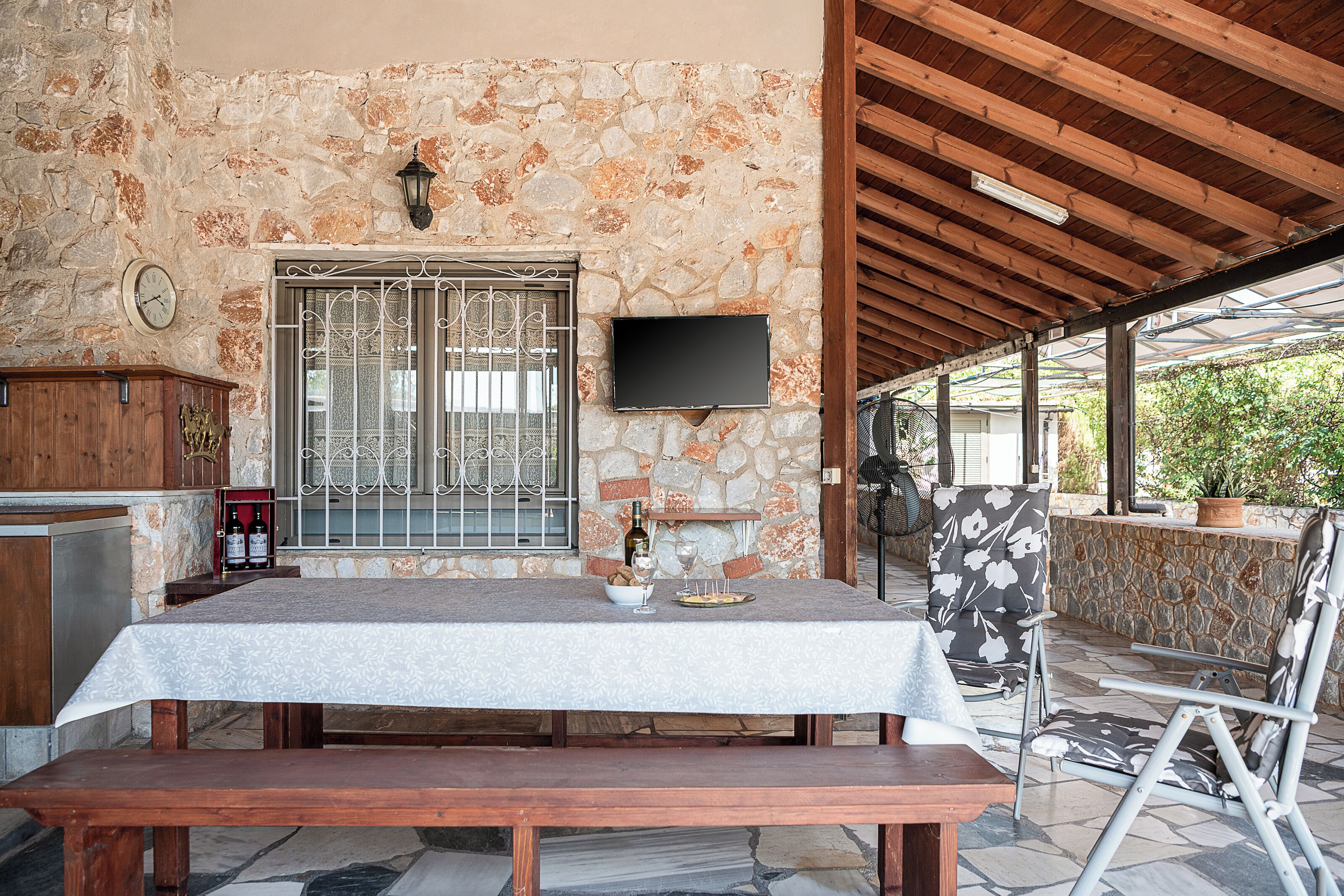 A stone-walled patio with a wooden table set for dining, a television mounted on the wall, and a bench.