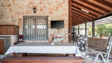 A stone-walled patio with a wooden table set for dining, a television mounted on the wall, and a bench.