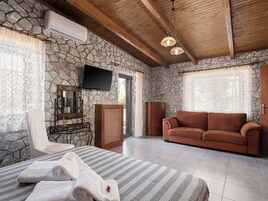 Una habitación con pared de piedra, techo de madera, televisor, sofá, sillón, cama con toallas y cómoda.