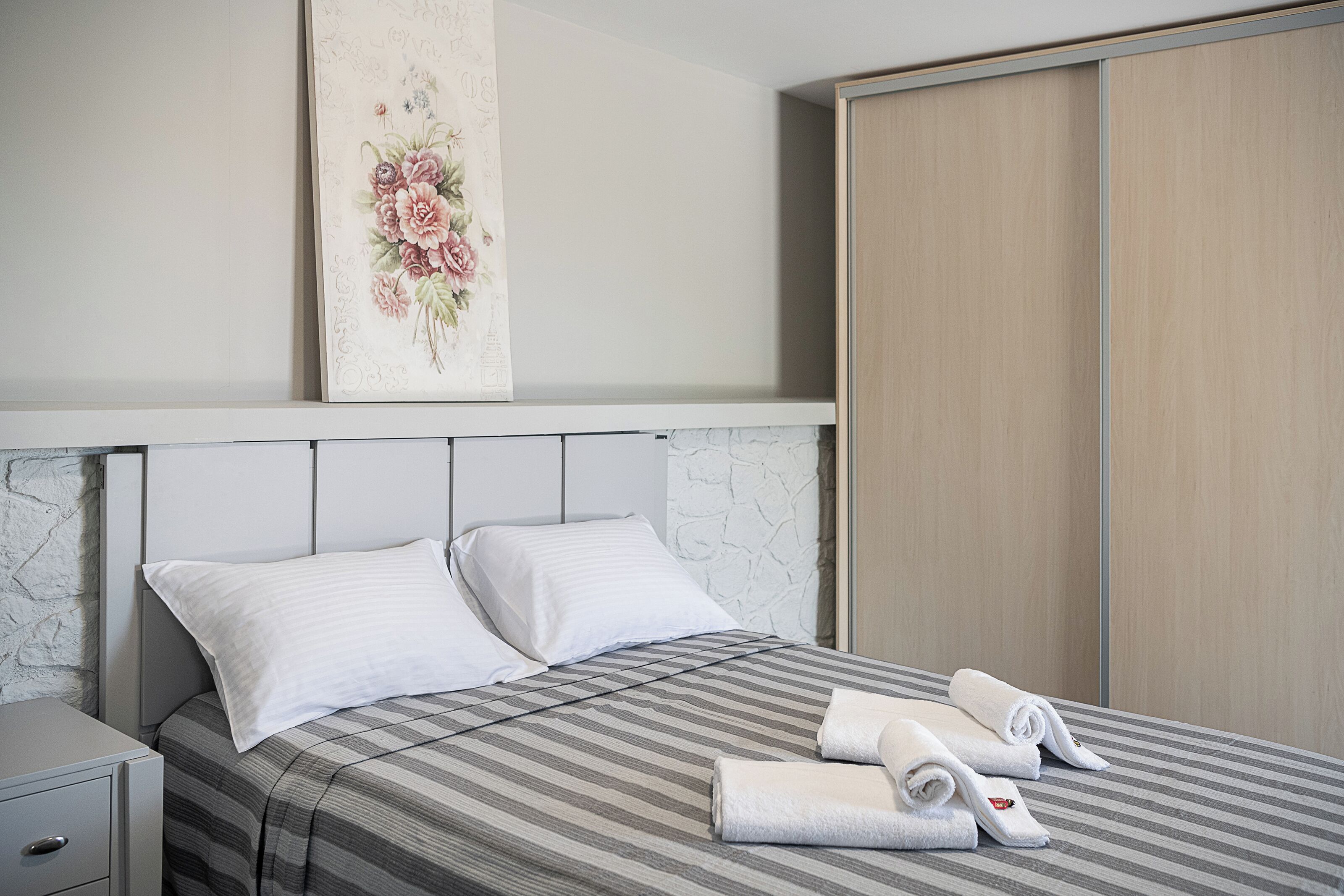 Una cama bien tendida con ropa de cama a rayas, dos almohadas y toallas enrolladas sobre una mesita de noche blanca.