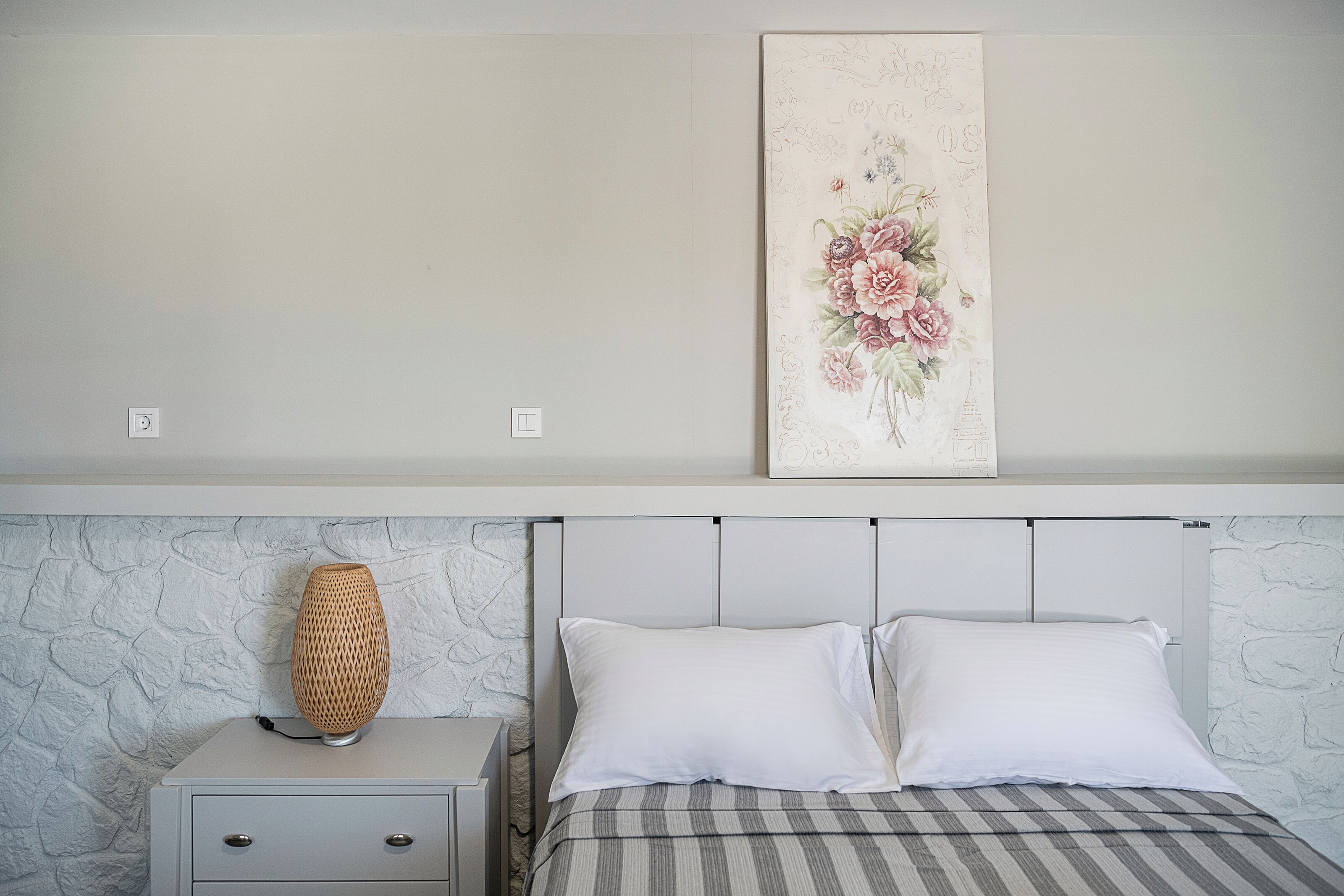 Un dormitorio con una cama, dos almohadas, una mesita de noche, una lámpara y una obra de arte floral en la pared.