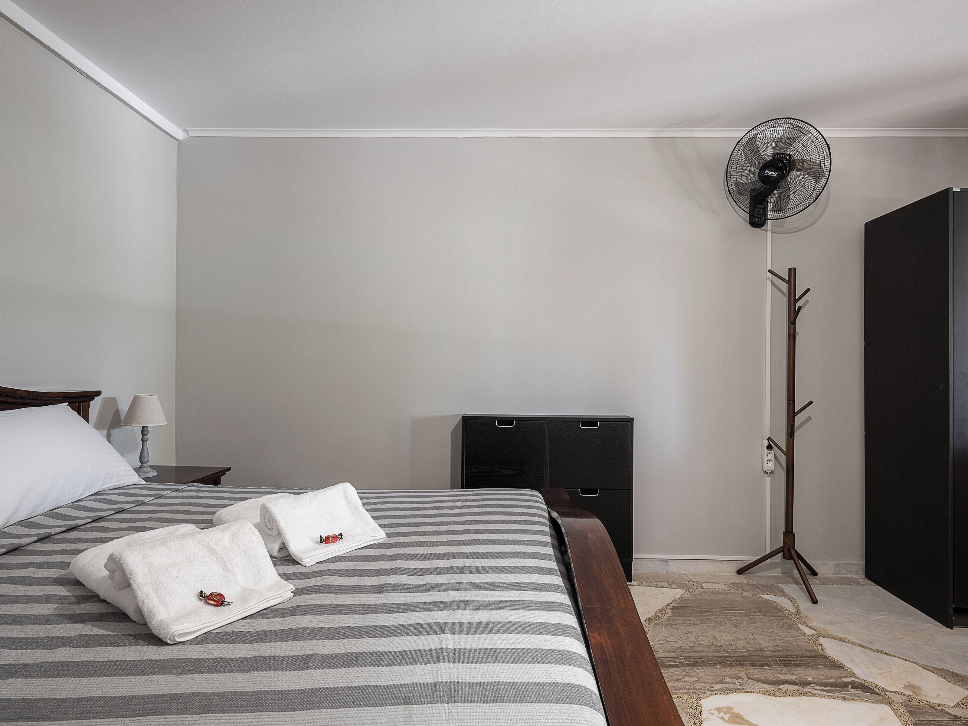 A neatly made bed with striped bedding and two towels, a standing fan, a coat rack, and a dark piece of furniture against a plain wall.