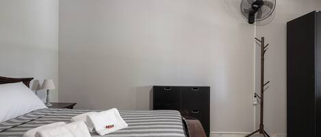 A neatly made bed with striped bedding and two towels, a standing fan, a coat rack, and a dark piece of furniture against a plain wall.
