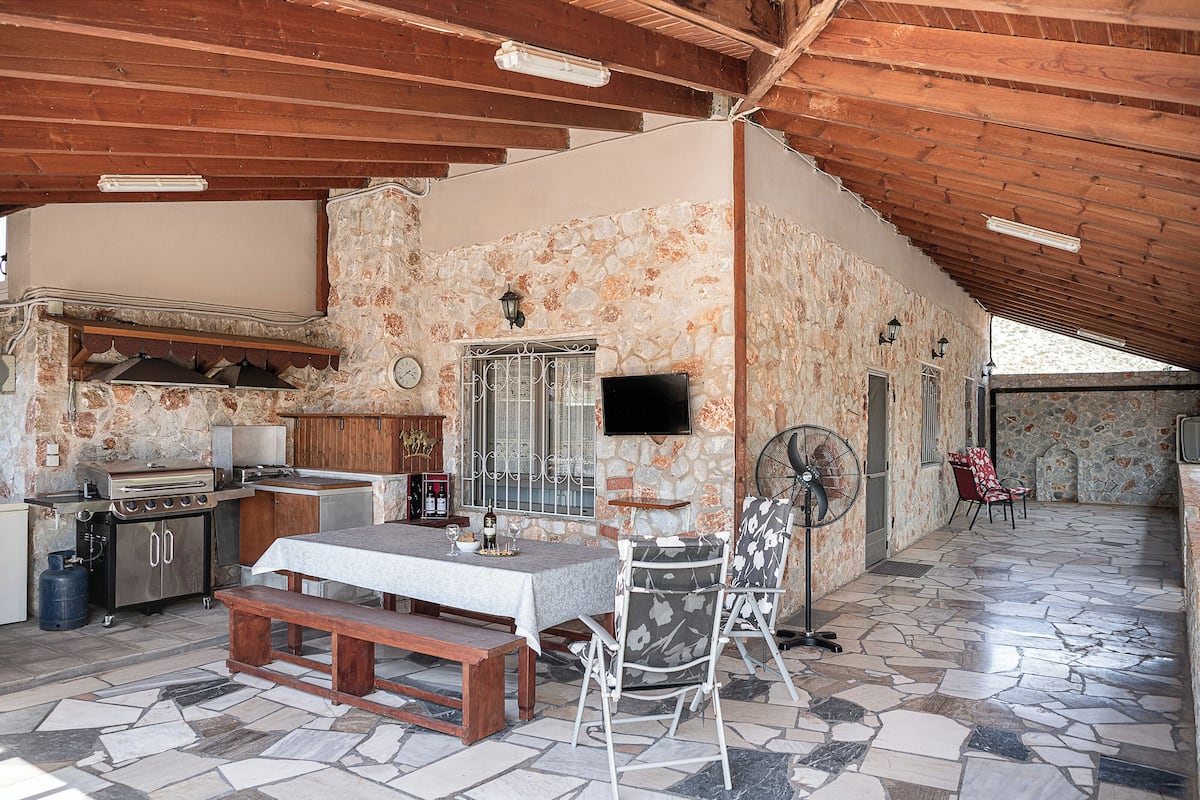 A rustic kitchen with stone walls, wooden beams, a dining table with benches, and a TV mounted on the wall.