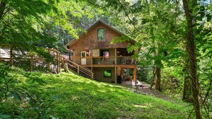 A two-story log cabin with a balcony, surrounded by dense forest. - On the Rocks: Riverside Relaxation Awaits (Sevierville)
