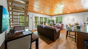 A spacious living area with a wooden ceiling, large windows, a sofa, a TV, and a dining area with a table and chairs. - On the Rocks: Riverside Relaxation Awaits (Sevierville)