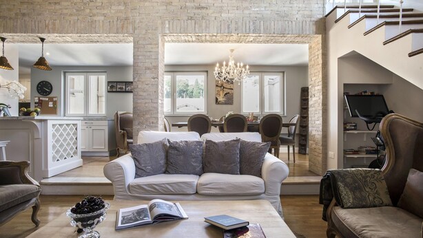 Un vaste espace de vie agrémenté d’un canapé blanc, d’une table basse avec des livres et d’un vase décoratif. On aperçoit à l’arrière-plan un coin repas avec un lustre.