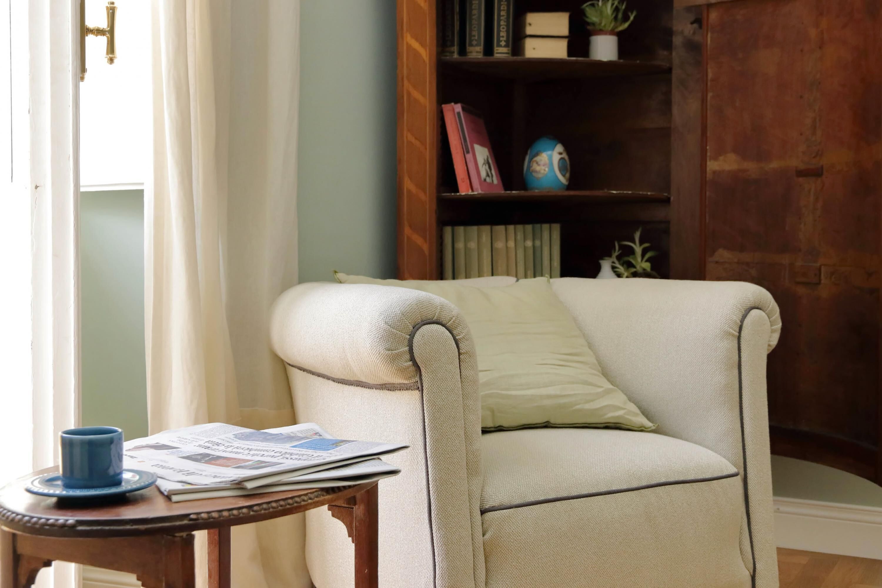 Une pièce comprenant un fauteuil de couleur claire, une petite table en bois avec une tasse bleue et des journaux, ainsi qu’une bibliothèque remplie de livres et d’objets décoratifs.