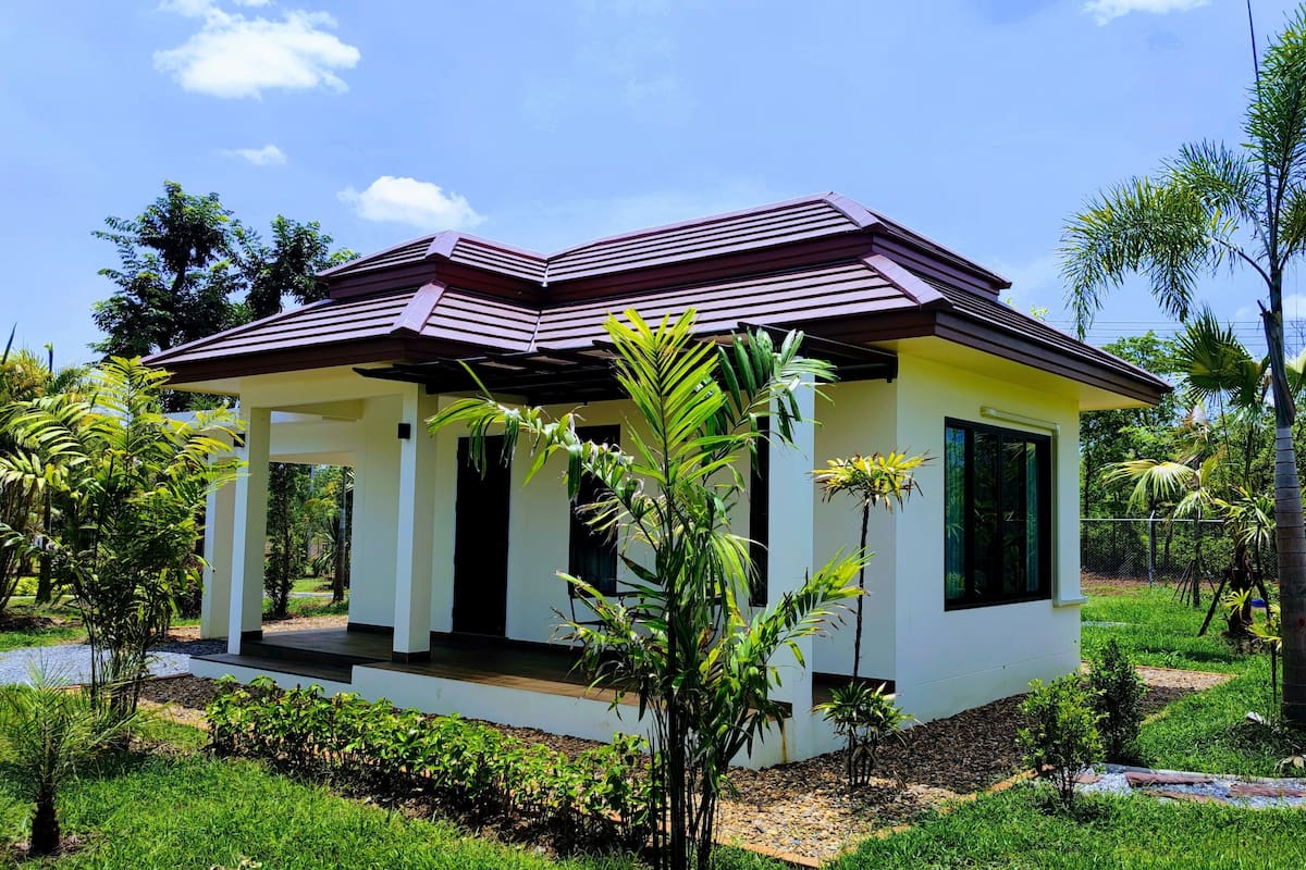 A small, single-story house with a red roof, surrounded by lush greenery and palm trees.
