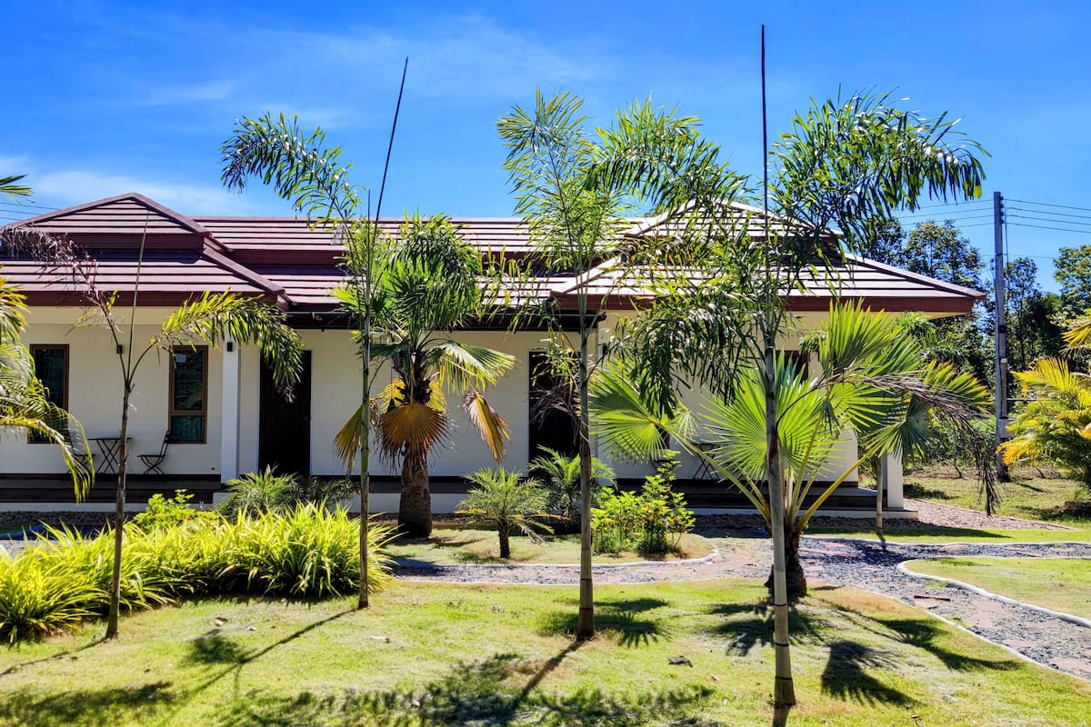 A two-story house with a red roof, surrounded by tropical vegetation and a clear blue sky.