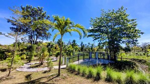 A bridge over a stone pathway surrounded by lush greenery and trees. - Isan Golf & Adventure Hotel (Phen)