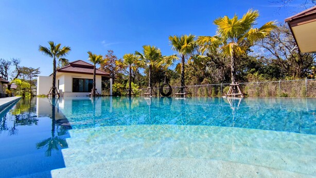 A clear swimming pool surrounded by palm trees and a building with a red roof. - Isan Golf & Adventure Hotel (Phen)