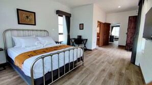 A neatly made bed with a metal frame, white bedding, and a brown decorative throw. There is a small table with a chair beside the bed, and a framed picture on the wall. The room has wooden flooring and a door leading to another room. - Isan Golf & Adventure Hotel (Phen)