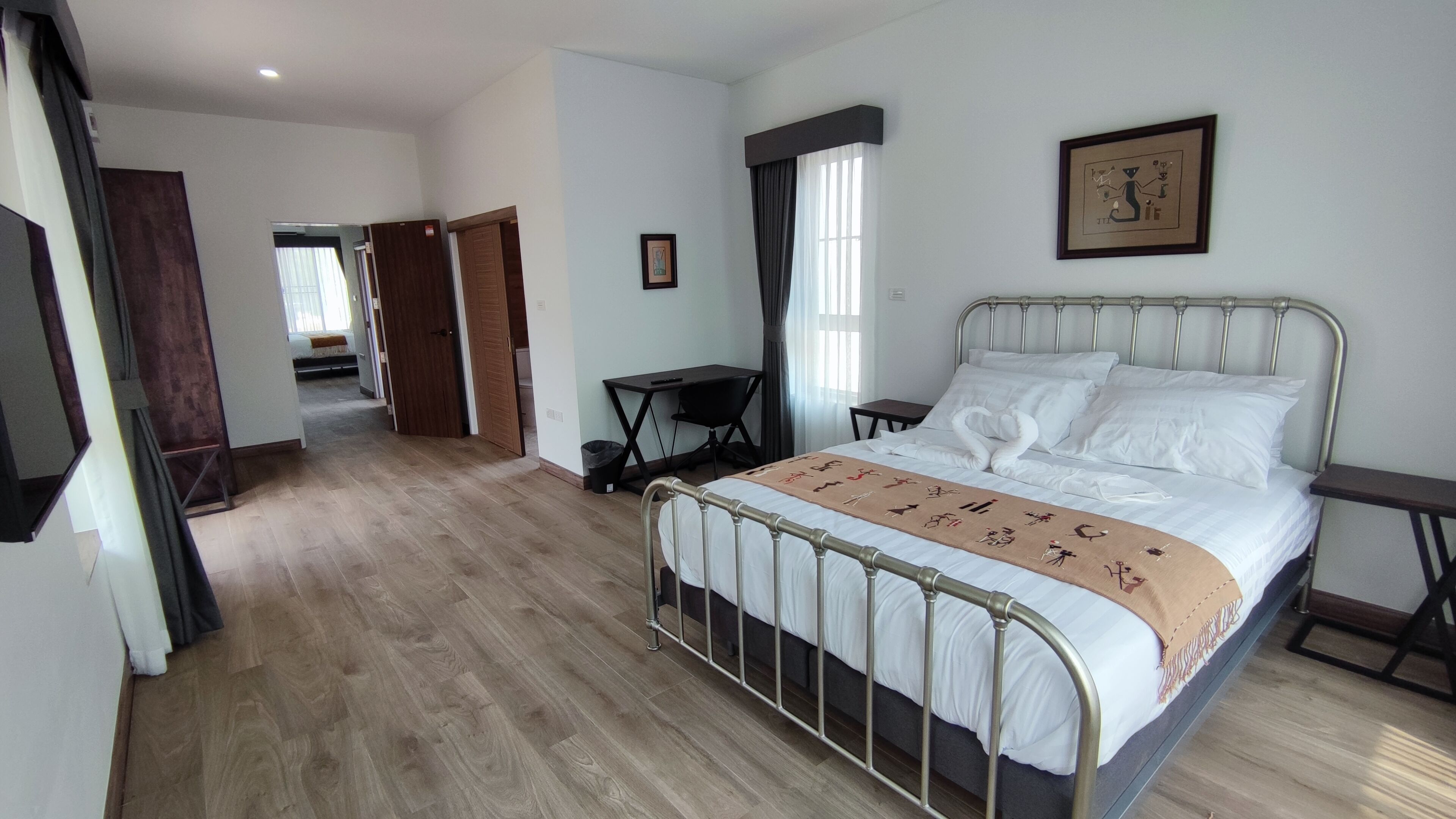 Un dormitorio con estructura de cama metálica, suelo de madera, un escritorio con silla y una ventana con cortinas.