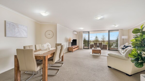 A modern living room with a dining area, a flat-screen TV, and a balcony with a view.